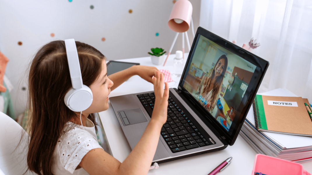 A young girl wearing headphones sits at a desk, waving at a woman on a laptop screen during a video call. A notebook, pen, and lamp are visible on the desk.