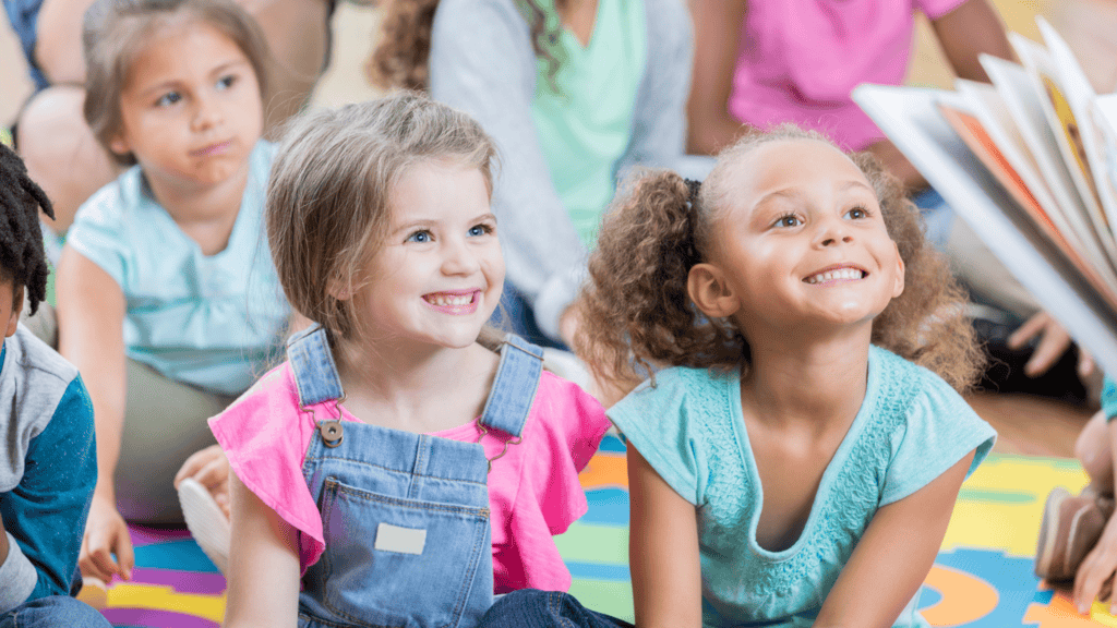 A group of young children sitting on a colorful mat in a classroom, smiling and looking at someone reading a book.