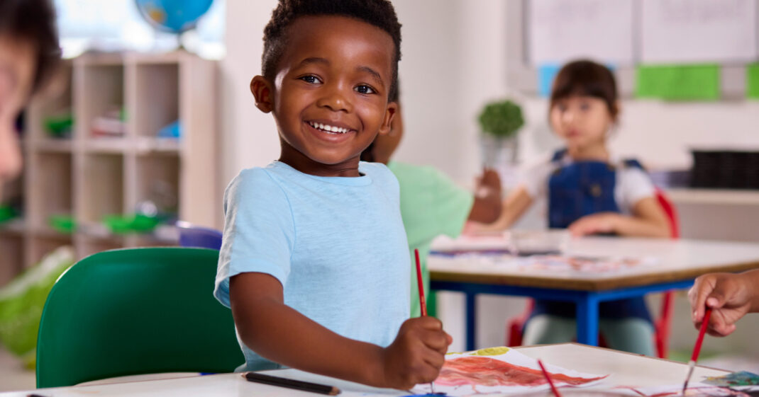 Smiling young boy sitting at a classroom table, holding a paintbrush and painting on paper, with other children in the background.