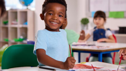 Smiling young boy sitting at a classroom table, holding a paintbrush and painting on paper, with other children in the background.