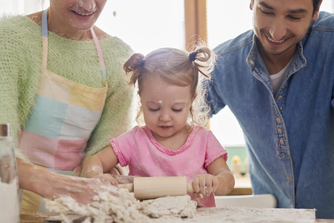 A young child uses a rolling pin on dough with the help of two adults, likely her parents, in a bright kitchen setting.